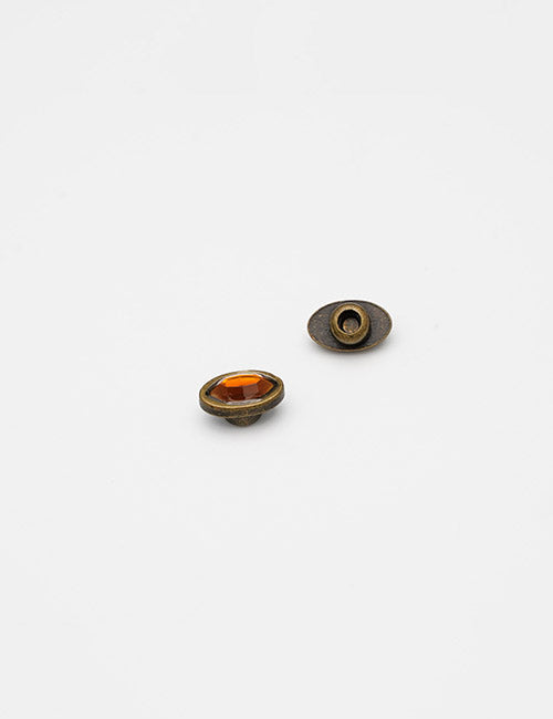 Two small, oval Adornments by YIPAI are displayed on a white background; one features an orange gem-like center with an antique metal finish, while the other shows its plain back.