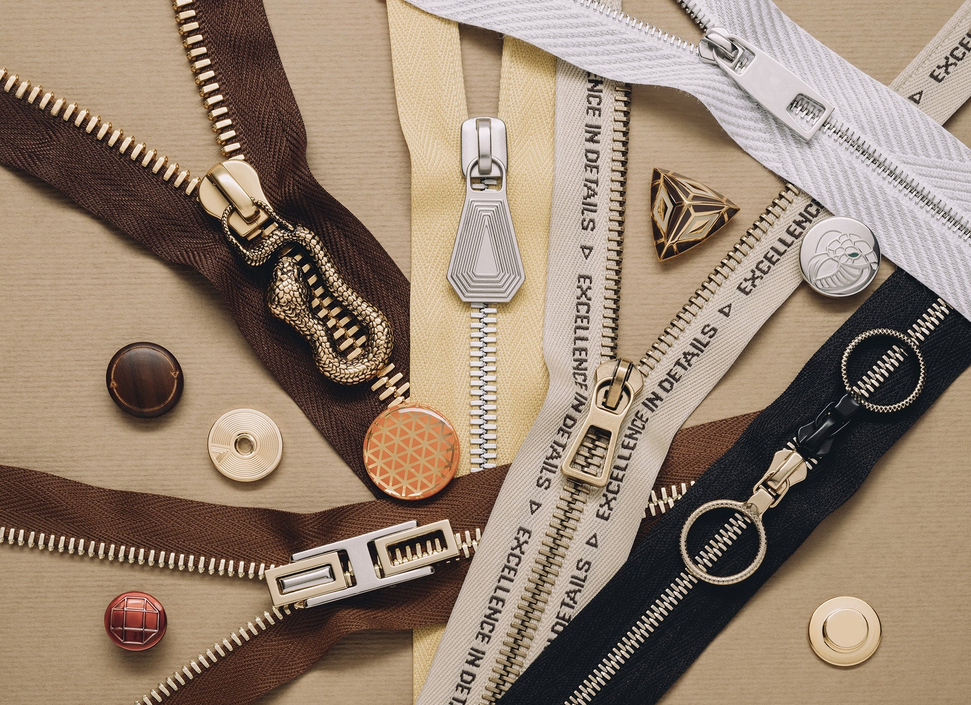 A variety of zippers in different colors, materials, and metallic finishes are arranged on a beige surface alongside several designer buttons and decorative embellishments.