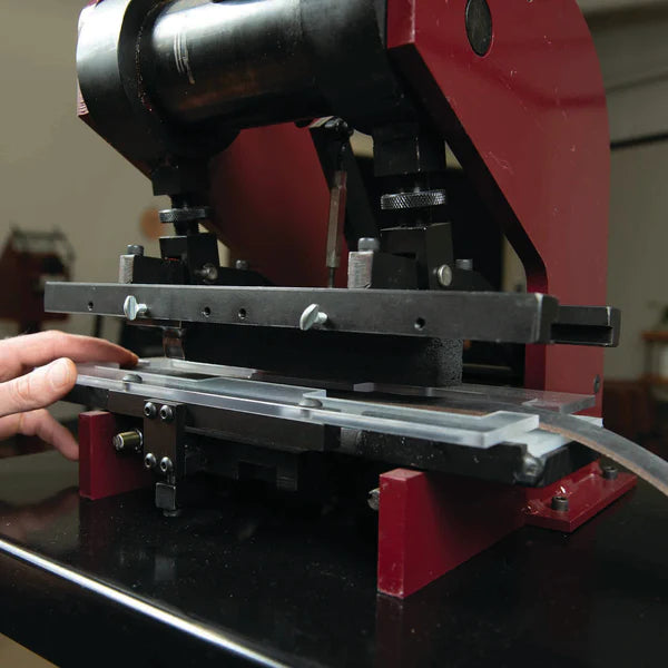 A close-up of a persons hand operating a red and black metal machine, possibly a press or cutting device, with a metal or plastic strip positioned under the tool.