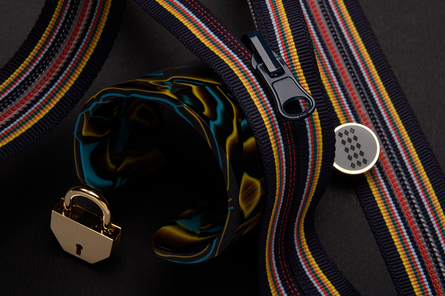 Colorful striped zippers, a marbled bracelet, a gold padlock, and a round pin with a grid pattern are arranged on a dark background.