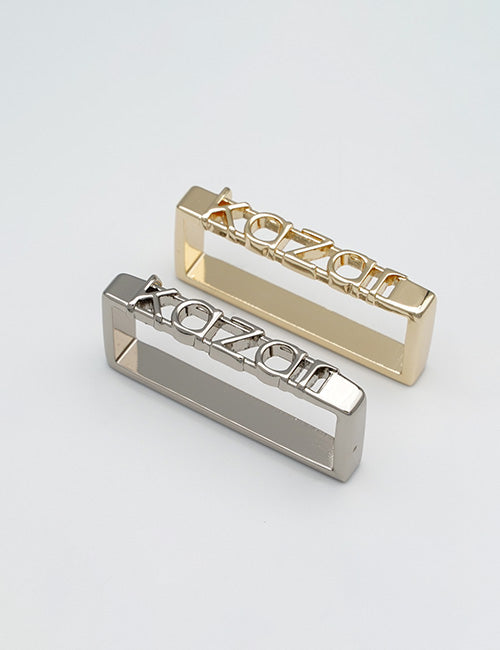 Two rectangular YIPAI Buckles, one gold and one silver, featuring the word KOZON cut out on top, are displayed on a plain white surface.