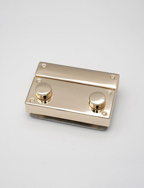 A shiny, rectangular YIPAI Closures gold metal object with two round knobs and four screws rests on a light gray surface.