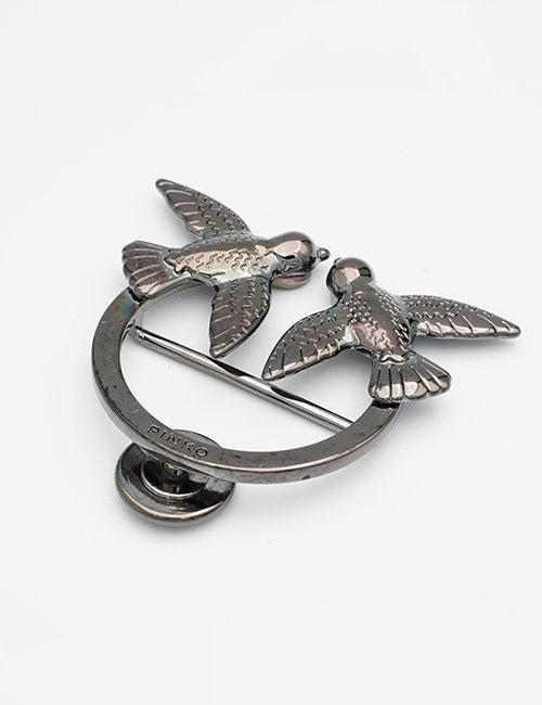 The YIPAI Closures brooch showcases two birds with outstretched wings facing each other, perched on a circular ring crossed by a pin, set against a plain white background.