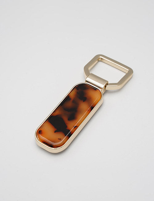 A YIPAI Fasteners gold keychain with a rectangular tortoiseshell-patterned accent rests on a plain light gray surface.