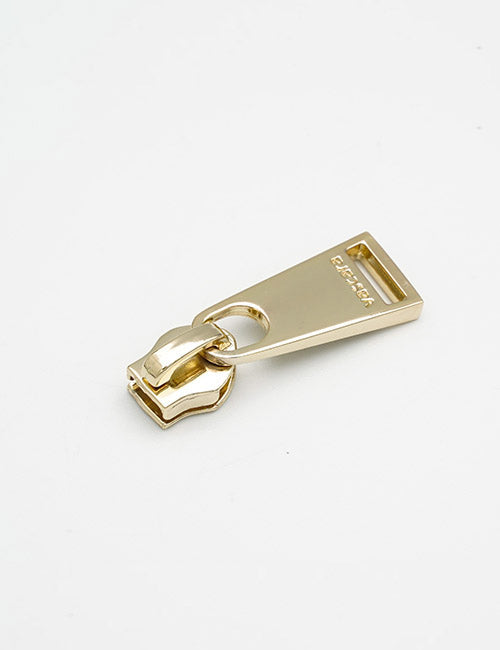 A shiny gold YIPAI Fasteners zipper pull tab with a rectangular hole at one end lies on a plain white surface. Excellence is engraved on the wide end of the tab.