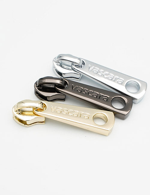 Three YIPAI Fasteners in silver, dark gray, and gold are arranged diagonally on a white background. Each metal zipper pull features the brand’s engraving.