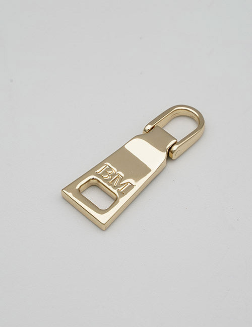 A gold-colored YIPAI Fasteners metal zipper pull tab rests on a light gray surface, with raised BM letters near the bottom.