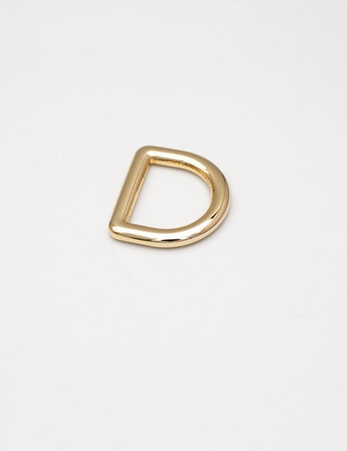 A YIPAI Rings & Slides gold D-ring metal buckle rests on a plain white surface, with its straight edge at the top and curved edge at the bottom.